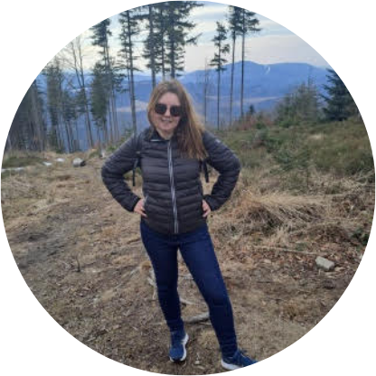 Osoba w kurtce puchowej pozuje na górskim szlaku w otoczeniu drzew, w tle widoczne góry.