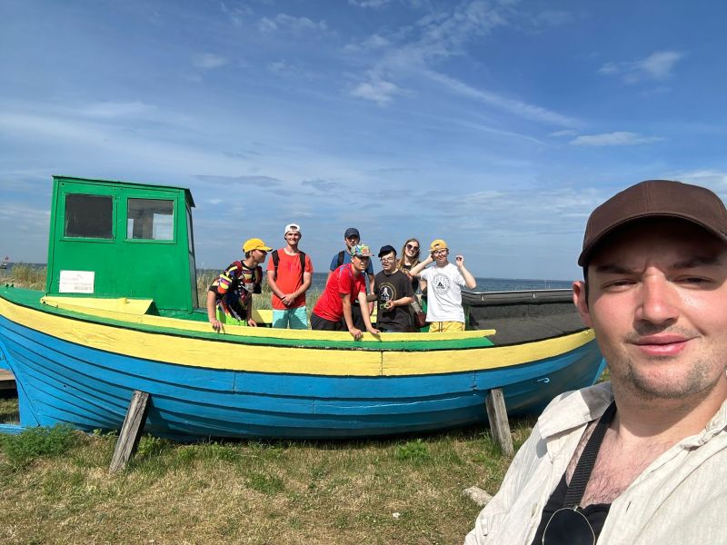 Grupa osób pozuje na niebiesko-żółtej łodzi, jedna robi selfie. W tle niebo i morze.