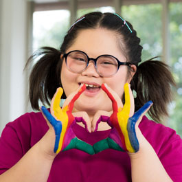 Osoba z uśmiechem i kolorowymi dłońmi układa znak serca, stojąc przed oknem.