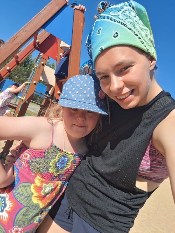 Dwie osoby uśmiechają się na placu zabaw, w jasny, słoneczny dzień. Noszą kolorowe chustki i kapelusz.