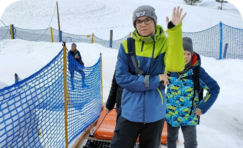 Osoba w zimowej odzieży na śniegu, macha ręką. W tle widoczna jest niebieska siatka i drzewa.