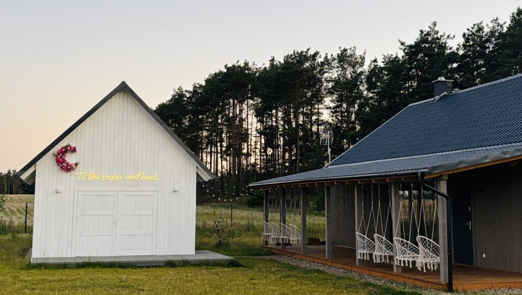 Biały domek z napisem To the moon and back pośród pól i lasów, obok altana z wiszącymi krzesłami.
