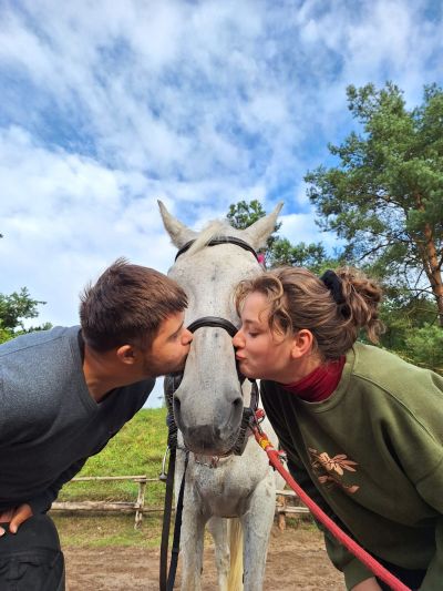 Dwie osoby całują konia po obu stronach jego pyska, stojąc na tle zieleni i nieba.