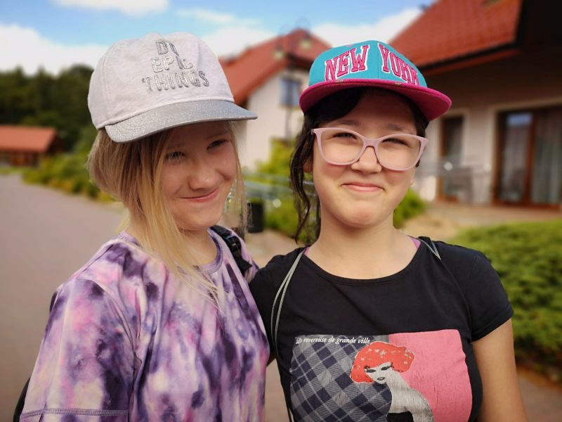 Dwie uśmiechnięte osoby w czapkach z daszkiem, na tle domów, w pogodny dzień.