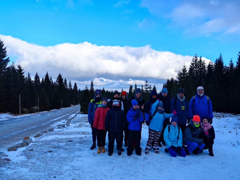 Grupa ludzi w zimowych ubraniach pozuje na ośnieżonej ścieżce w górach, otoczona drzewami i niebem z chmurami.