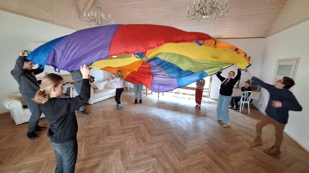 Grupa dzieci bawi się kolorowym spadochronem w przestronnym, jasnym pomieszczeniu z drewnianą podłogą.