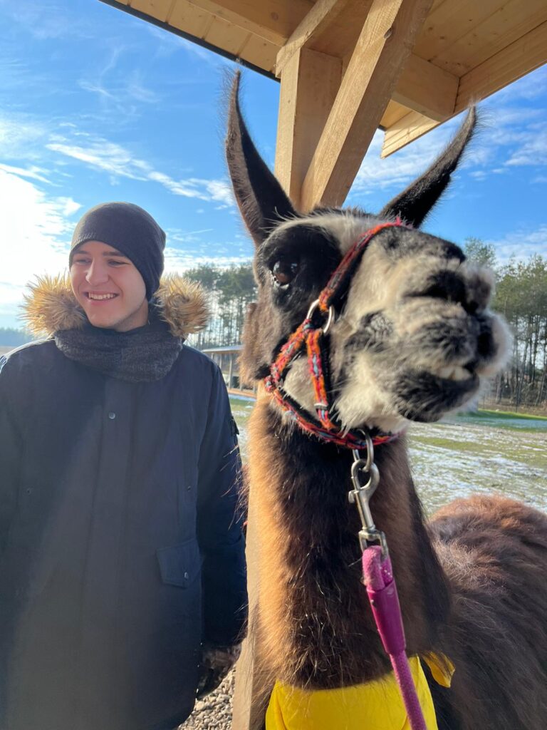 Uśmiechnięta osoba w czapce zimowej i lama z kolorową uprzężą stoją obok siebie przy drewnianej altanie.