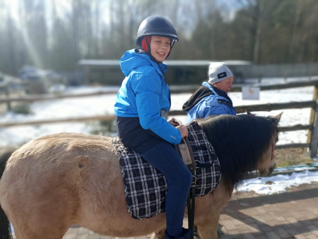 Chłopiec na koniu w niebieskiej kurtce i kasku, uśmiecha się, a w tle zima i zaplecze gospodarstwa.