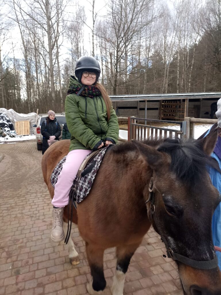 Osoba w zielonej kurtce i kasku siedzi na koniu na brukowanej drodze, w tle zimowy las i budynki.