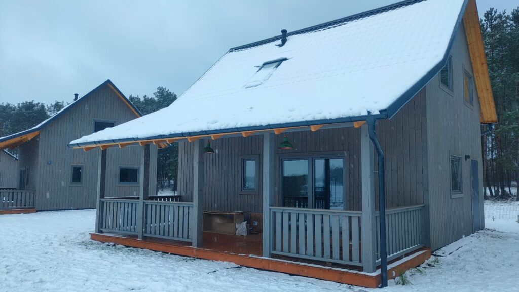 Dwa drewniane domki pokryte śniegiem na tle lasu zimą, z zadaszonymi werandami.