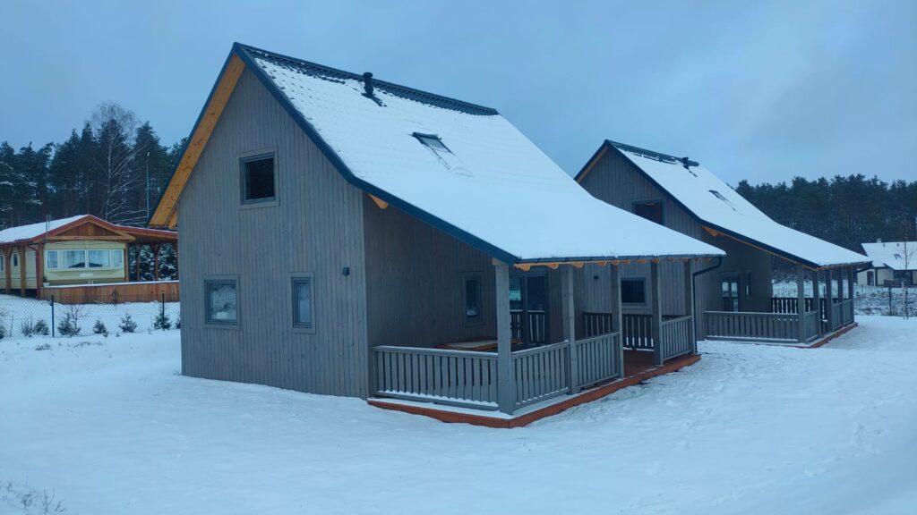 Dwa szare drewniane domki z zadaszonymi werandami, pokryte śniegiem, na tle lasu zimą.