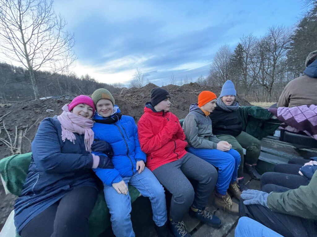 Grupa uśmiechniętych osób siedzi na otwartej przyczepie w zimowych ubraniach, na tle drzew i ziemi.