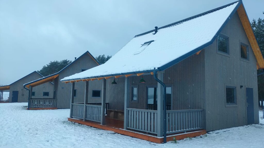 Drewniane domki przysypane śniegiem, z zadaszonymi werandami na tle chmur i drzew.