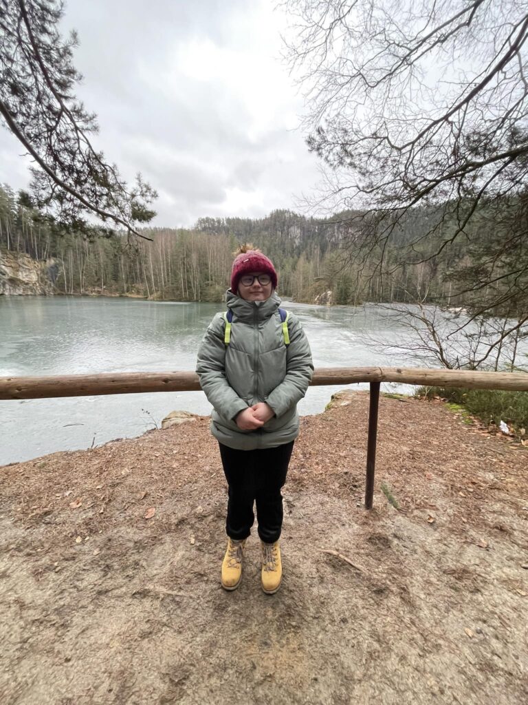 Osoba w zimowym ubraniu stoi przy jeziorze w otoczeniu drzew i gór. Pochmurne niebo.