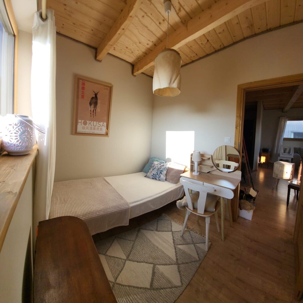 Przytulny pokój z łóżkiem, biurkiem i lampą. Na ścianie plakat, na podłodze dywanik. Drewniany sufit i podłoga.