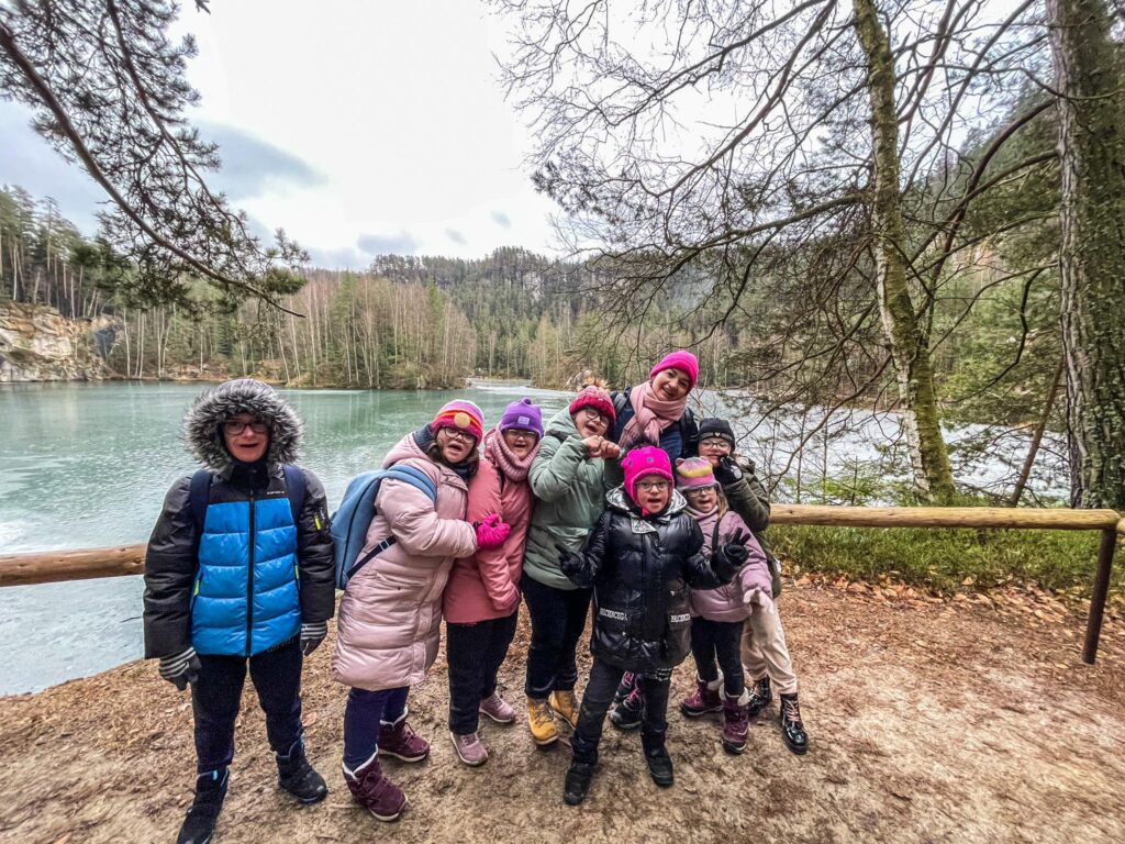 Grupa ludzi w zimowych ubraniach pozuje nad jeziorem w otoczeniu lasu, uśmiechy na twarzach.