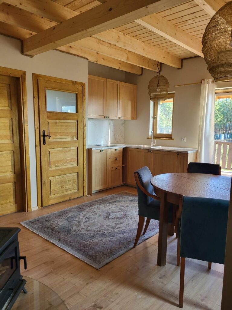 Przytulna kuchnia w drewnie z okrągłym stołem, dywanem, jasną lampą i widokiem na las za oknem.