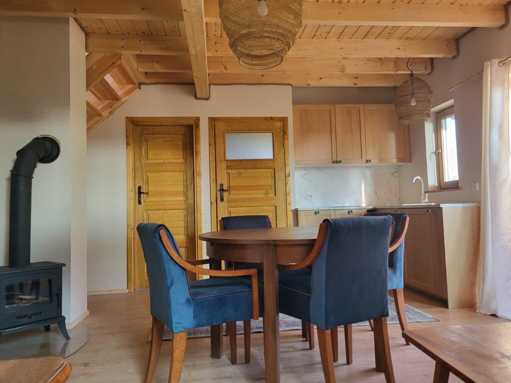 Drewniane wnętrze z jadalnią i kuchnią. Niebieskie krzesła, owalny stół, drewniane szafki i piecyk na lewo.