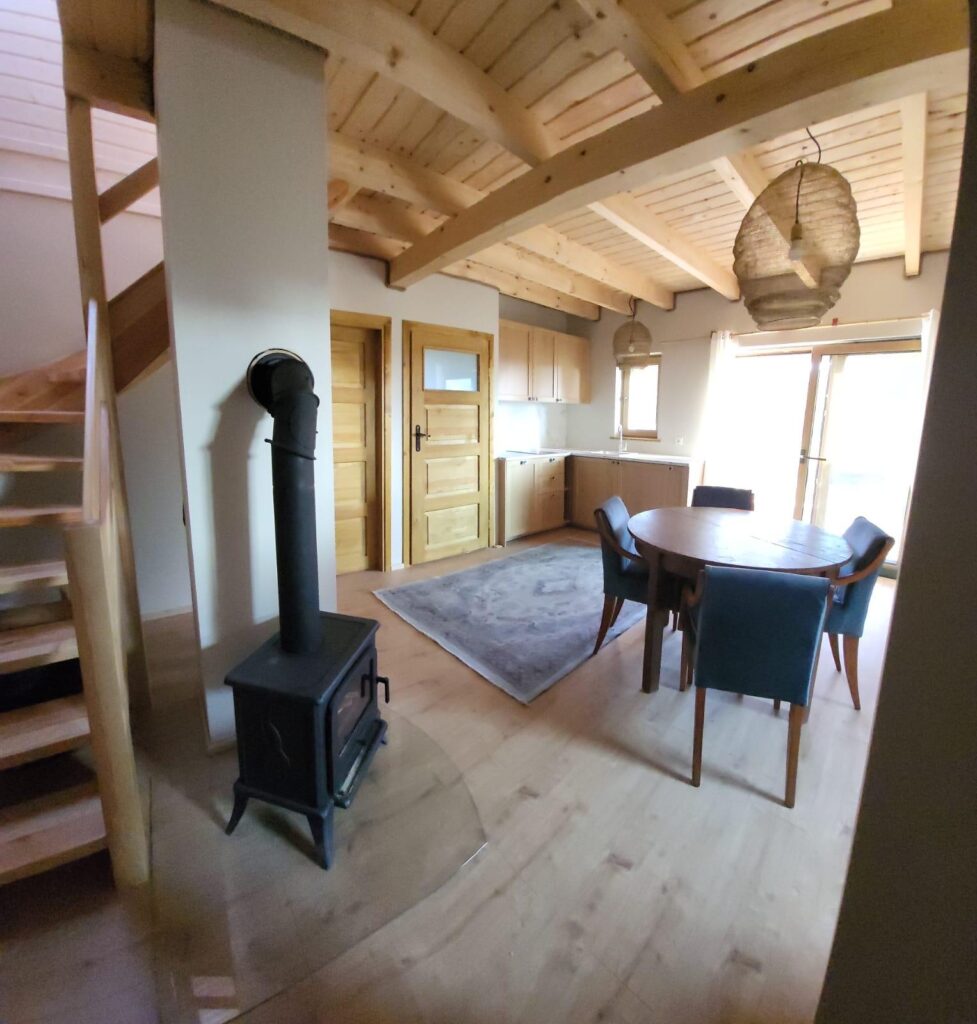 Przytulne wnętrze z drewnianym sufitem, piecem na drewno, okrągłym stołem i krzesłami, w tle schody.