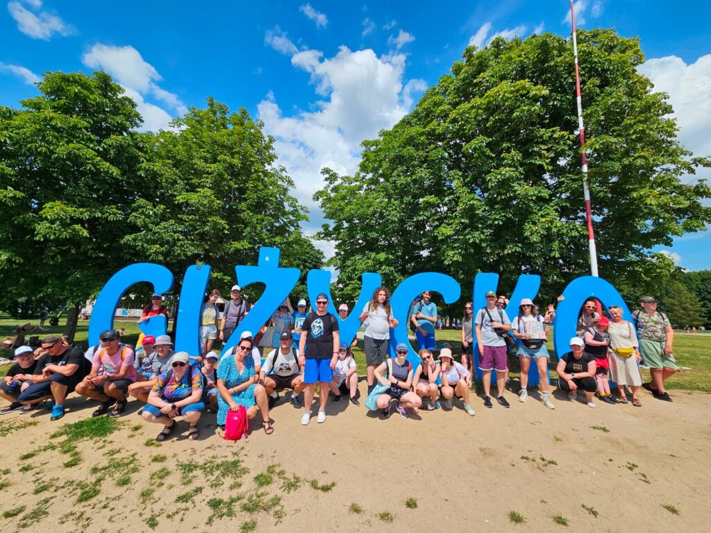 Grupa ludzi pozuje przed dużym, niebieskim napisem GIŻYCKO na tle zielonych drzew i błękitnego nieba.