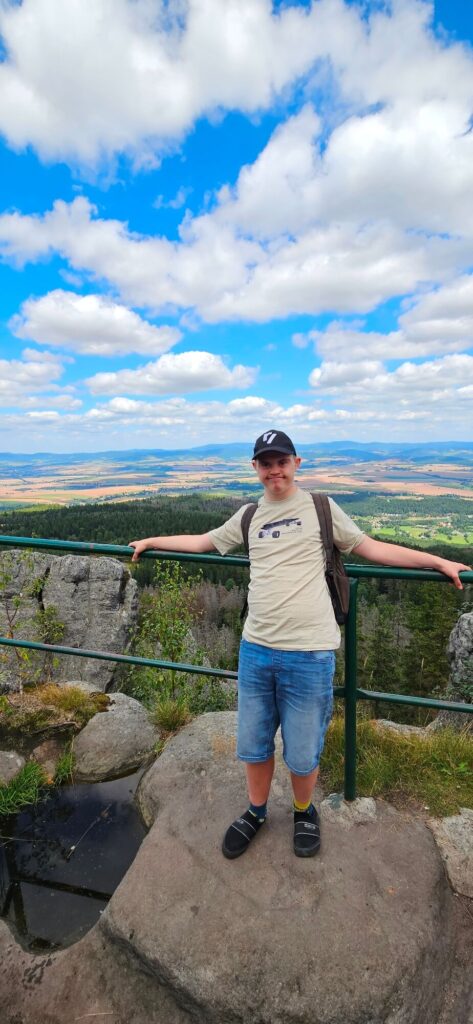 Osoba na szczycie góry przy poręczy, w tle rozległa panorama doliny i niebo z chmurami.