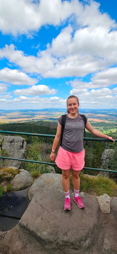 Osoba w różowych spodenkach stoi na skale z plecakiem, w tle rozległy krajobraz i niebo z chmurami.