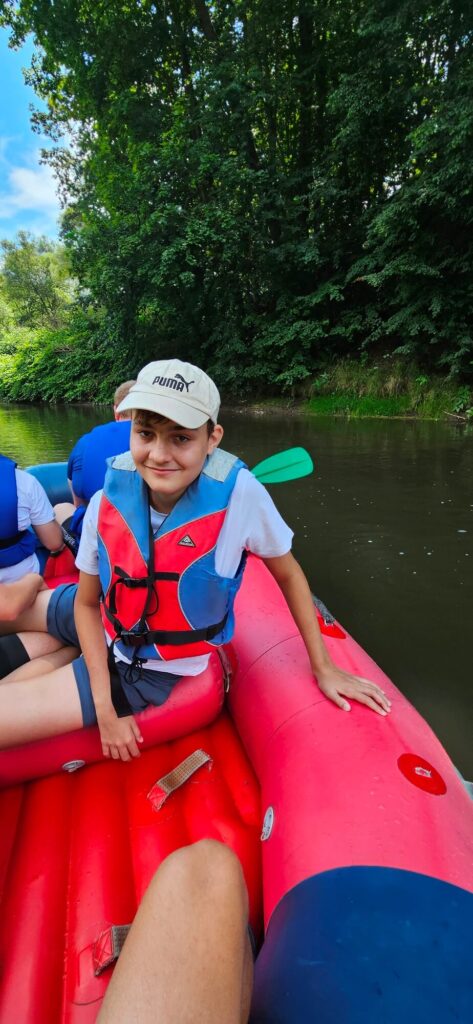 Osoba w kamizelce ratunkowej siedzi w czerwonym pontonie na rzece, na tle zielonego lasu.