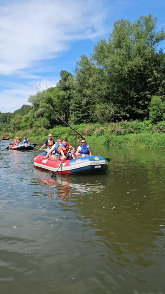 Grupa osób na spływie pontonowym po rzece, otoczona zielonymi drzewami i błękitnym niebem.