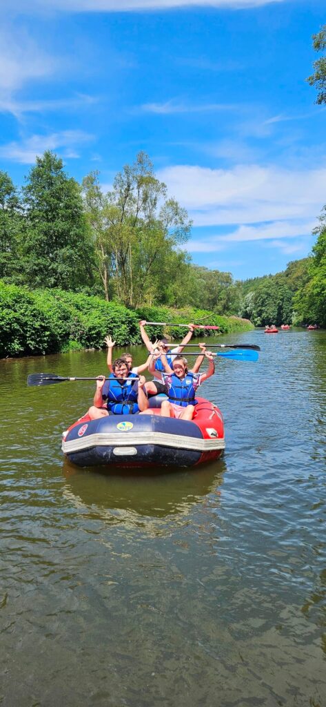 Grupa osób na pontonie wiosłująca na rzece, w otoczeniu zielonych drzew, w letni, słoneczny dzień.