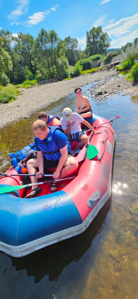Grupa ludzi w pontonie na płytkiej rzece, w otoczeniu zieleni i błękitnego nieba.