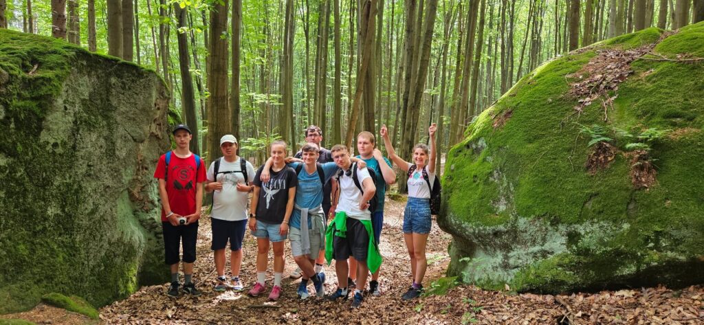 Grupa młodych osób na wycieczce w lesie, stojąca pomiędzy dwoma dużymi, zielonymi skałami porośniętymi mchem.