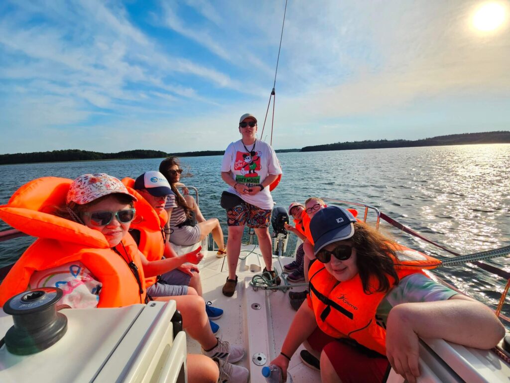 Grupa osób na jachcie w pogodny dzień, część w pomarańczowych kamizelkach ratunkowych, w tle spokojna woda i niebo.