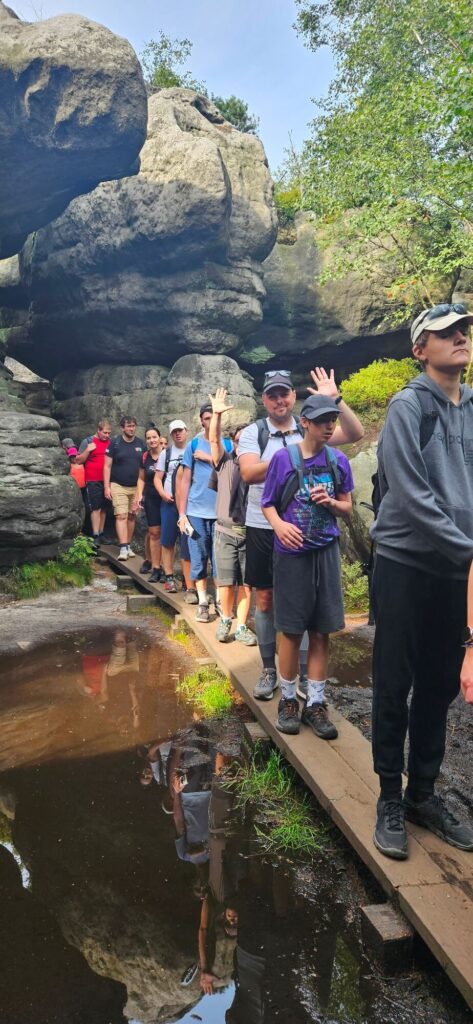 Grupa ludzi stoi w kolejce na drewnianej kładce pomiędzy skałami, niektórzy machają do kamery.