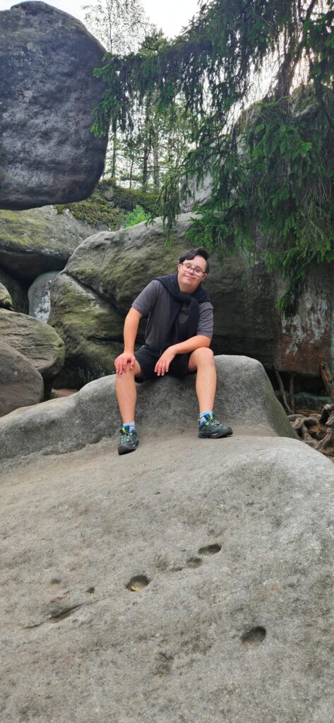 Osoba siedząca na dużym kamieniu wśród skał i drzew, uśmiechnięta, w casualowym stroju, w tle zielone drzewa.