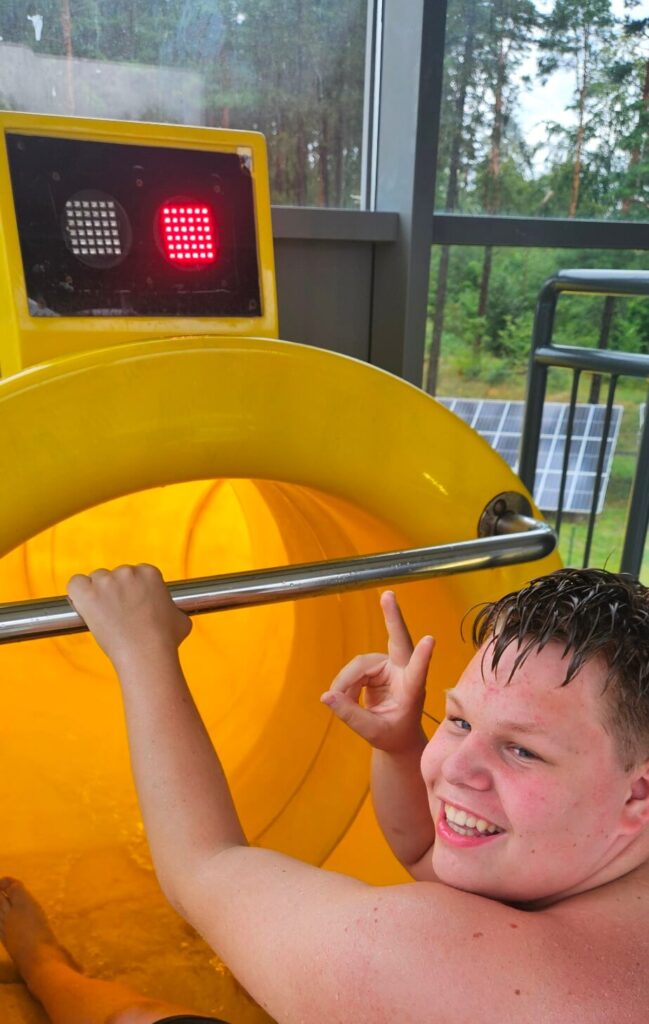 Osoba uśmiecha się przed żółtą zjeżdżalnią wodną z czerwonym światłem w tle, wskazując znak pokoju.