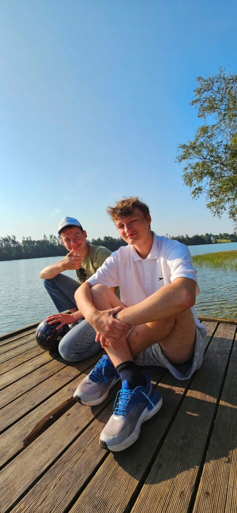 Dwóch mężczyzn siedzi na drewnianym pomoście nad jeziorem w słoneczny dzień, obaj uśmiechnięci i zrelaksowani.