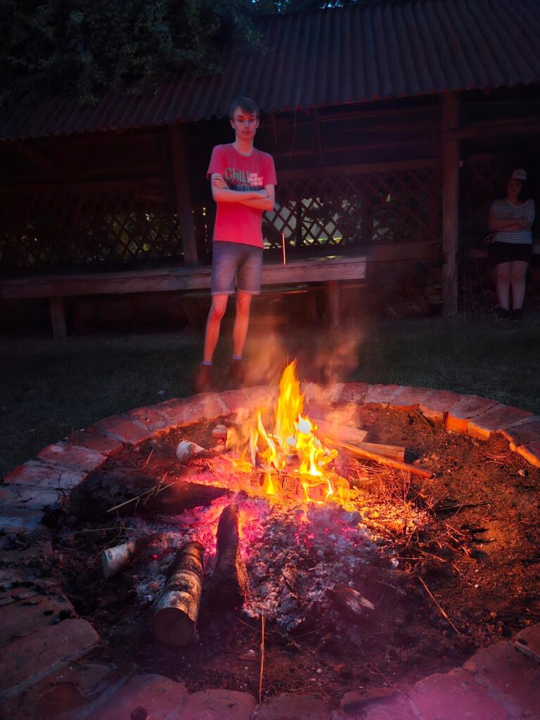 Osoba stoi przy ognisku w letni wieczór, w tle drewniana altana, atmosfera relaksu i ciepła.