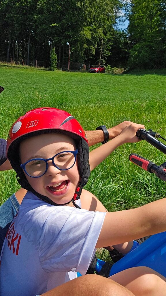 Dziecko w czerwonym kasku i okularach uśmiecha się, siedząc na rowerze w zielonym otoczeniu na tle lasu.