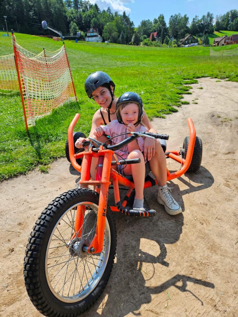 Dwie osoby w kaskach siedzące na pomarańczowym pojeździe terenowym na tle zielonego wzgórza w pogodny dzień.