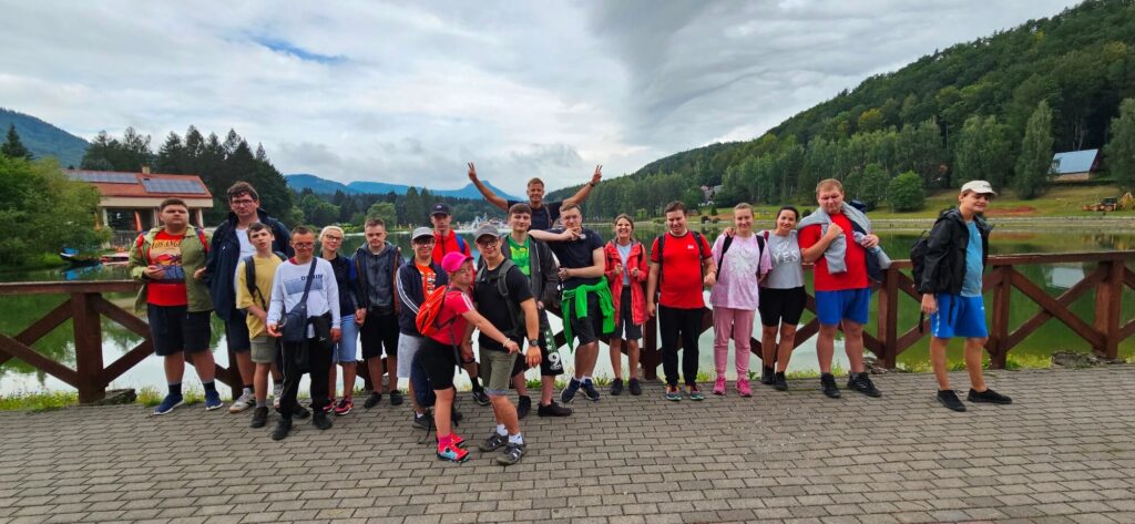 Grupa osób pozuje do zdjęcia na tle jeziora i drzew, słońce nad nimi skrywa się za chmurami.