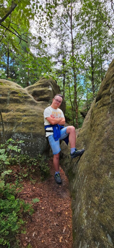 Osoba stojąca w lesie między dużymi skałami, z rękami skrzyżowanymi na piersi, w letnich ubraniach.