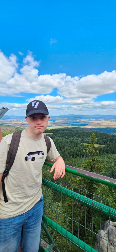 Osoba w czapce i plecaku stoi na tarasie widokowym z rozległym widokiem na góry i lasy.