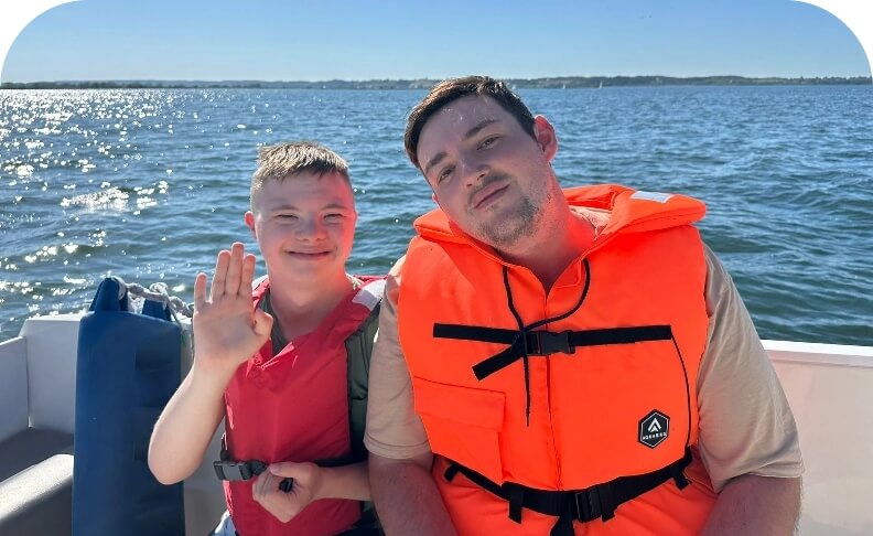 Dwóch mężczyzn w kamizelkach ratunkowych na łodzi, jeden z nich macha. W tle widać jezioro i pogodny dzień.