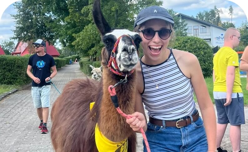 Kobieta w okularach przeciwsłonecznych przytula lamę na spacerze w parku; w tle inni spacerowicze.