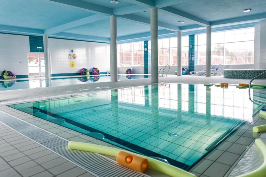 Wnętrze basenu krytego z jasną wodą, sprzętem do pływania i leżakami przy oknach.