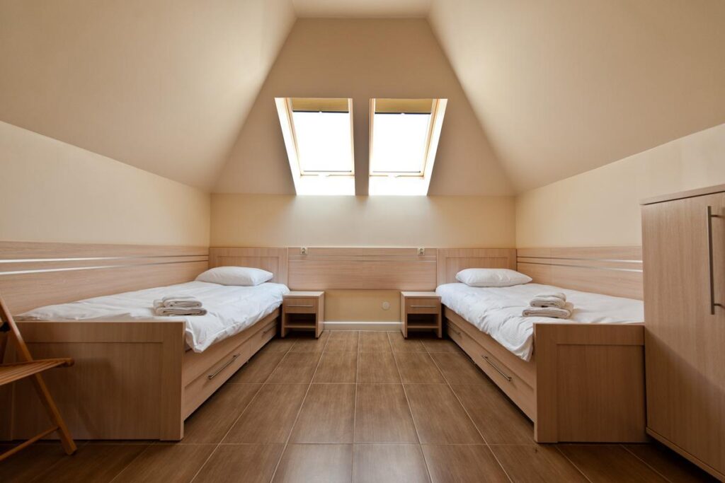 Dwa pojedyncze łóżka w jasnym pokoju z oknem dachowym, drewniane meble i ręczniki na łóżkach.