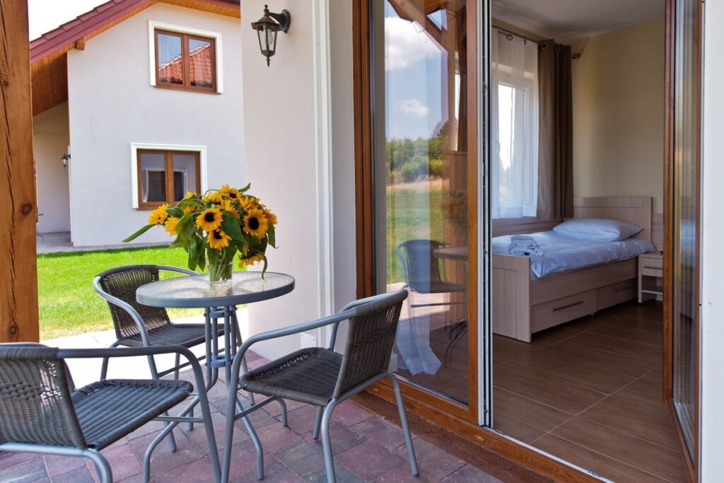 Widok tarasu z okrągłym stolikiem i krzesłami, wazon słoneczników, otwarte drzwi do przytulnego, jasnego pokoju.