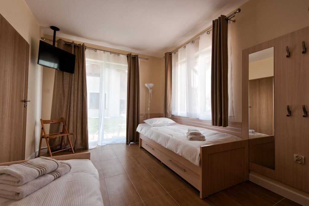 Pokój z łóżkami, telewizorem, lustrem i balkonem. Przytulny, jasny wystrój z brązowymi zasłonami.