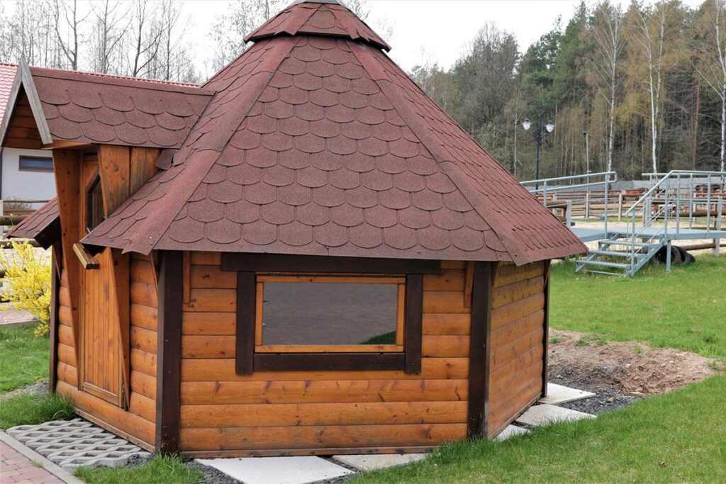 Drewniana altanka z oktagonalnym dachem, stojąca na trawniku, otoczona drzewami i ścieżką z kostki brukowej.