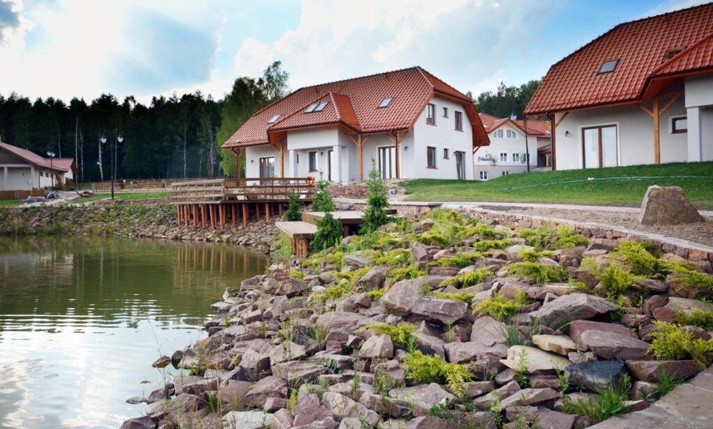 Nowoczesne domy z czerwonymi dachami nad brzegiem stawu, otoczone zielenią i drzewami w tle.